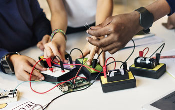 one hand holding tweezers while 2 others building electronics.  36 black and red wires plugged in to boards