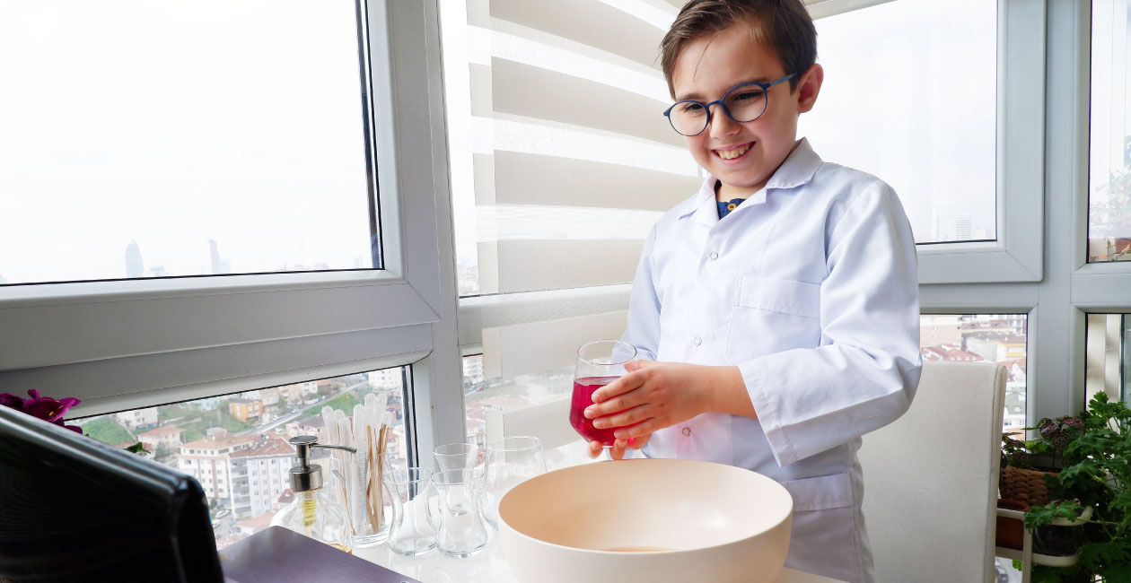 Boy in lab coat holding a glass with a red liquid standing in front of his computer performing a Mad Science virtual class.